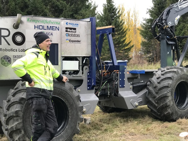 Man i gul jacka står vid skogsmaskin. Foto.