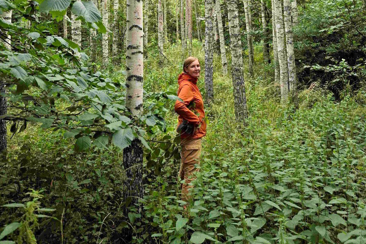 Kvinna i orange jacka står i lövskog. Foto.